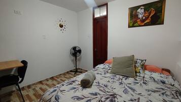 A bedroom with a bed, a desk, a chair, a standing fan, and a door.