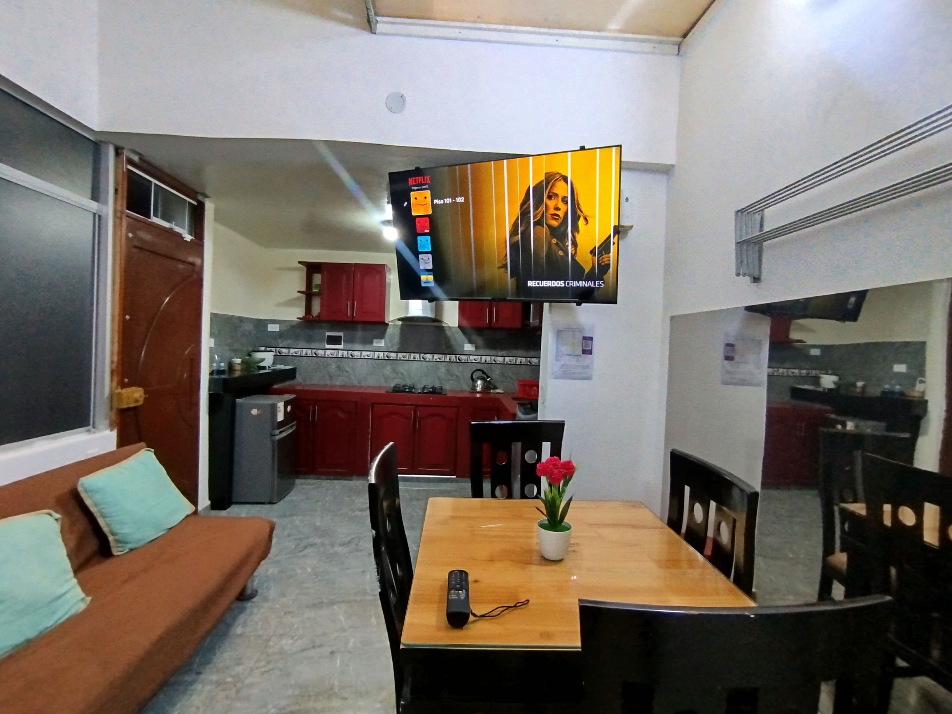 Un espacio habitable compacto con un sofá, una mesa de comedor con sillas, un televisor en la pared y una kitchenette al fondo.