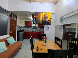 A compact living space with a sofa, a dining table with chairs, a TV mounted on the wall, and a kitchenette in the background.