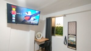 A room with a television mounted on the wall, a desk with a chair, a window with a view of greenery, and a wall-mounted fan. - Departamentos Appart d'Eliette (Trujillo)