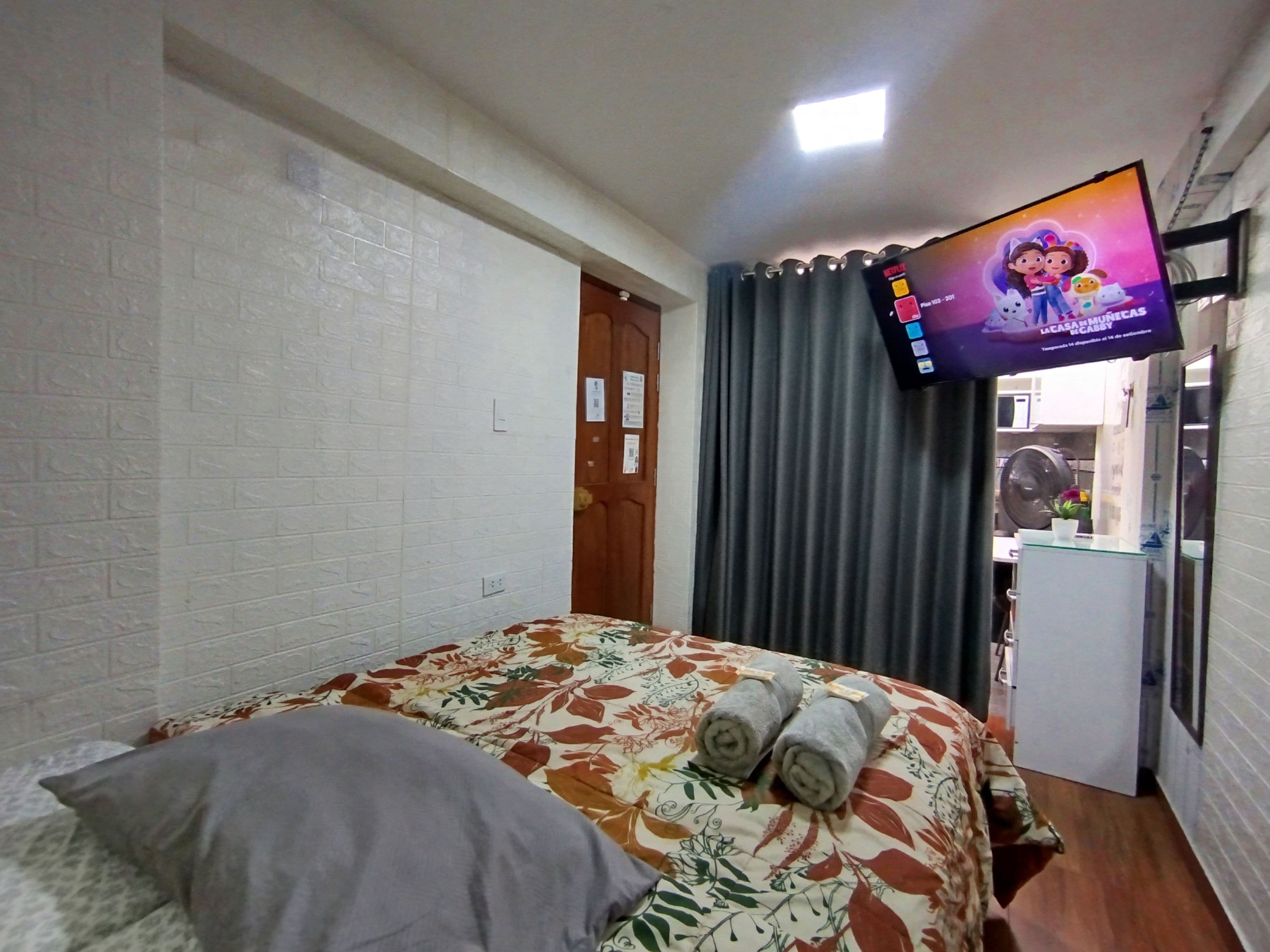 A hotel room with a bed, a television mounted on the wall, a door, and a small kitchenette area.