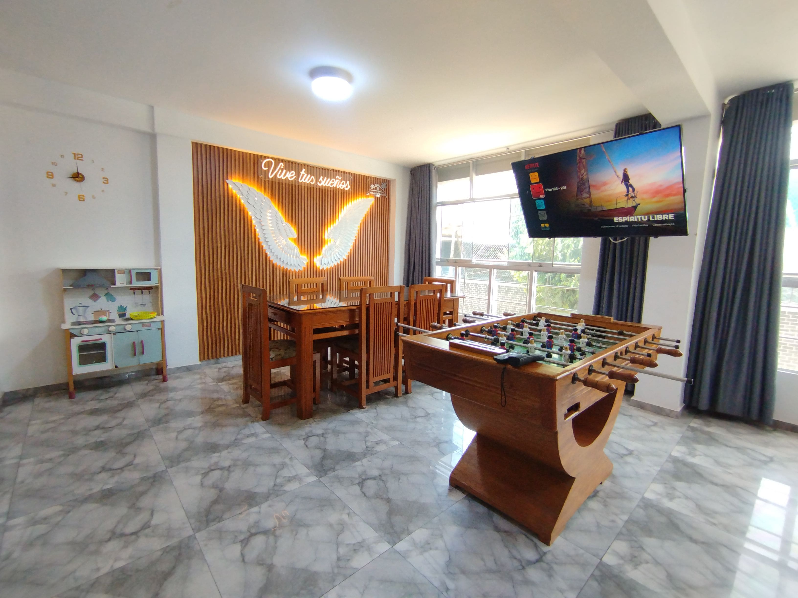 Quarto com mesa de pebolim, televisão, área de cozinha e parede com iluminação decorativa.