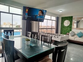 Una sala de estar moderna con una mesa de comedor de vidrio, mesa de fútbol, televisor montado en la pared y un adorno de pared verde.