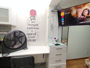 Quarto com escrivaninha, relógio de parede, televisão de tela plana, ventilador e parede decorativa com uma citação.