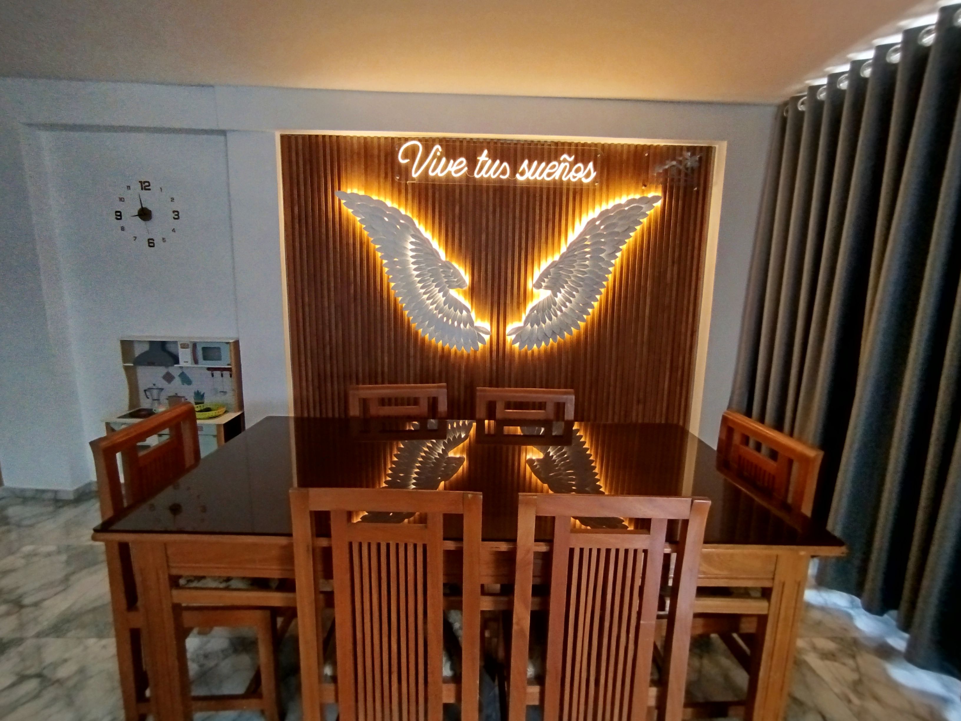 A dining area with a wooden table and chairs, a neon sign with wings, and a clock on the wall.