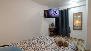 Habitación de hotel con una cama, televisión montada en la pared, escritorio con silla y un espejo.