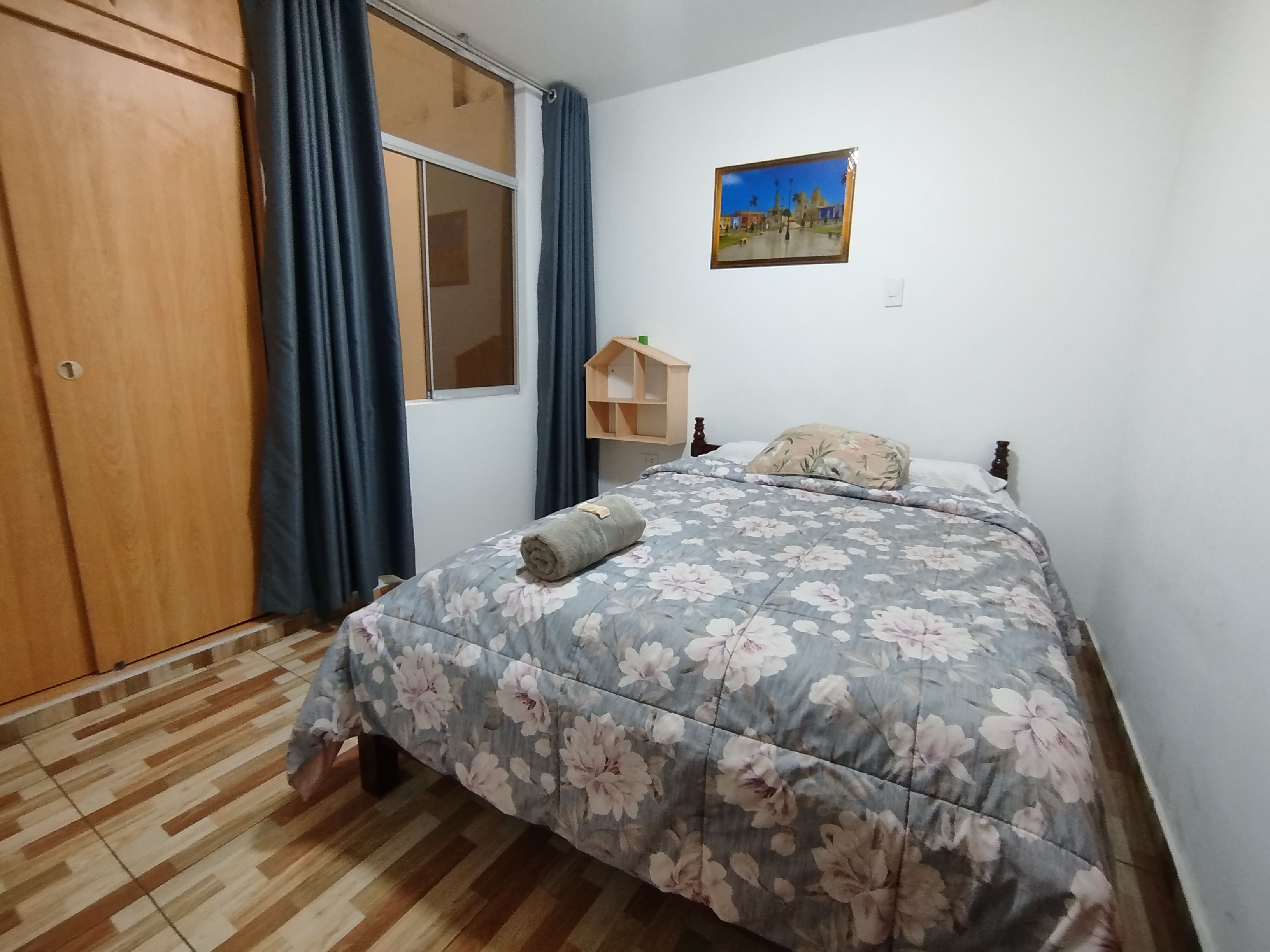 Un dormitorio con un armario de madera, una cama con edredón floral, una ventana con cortinas y un cuadro colgado en la pared.