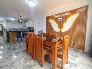 Un comedor con mesa y sillas de madera, una cocina al fondo y una pared con lámpara decorativa y un cartel.