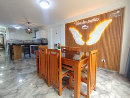 Un comedor con mesa y sillas de madera, una cocina al fondo y una pared con lámpara decorativa y un cartel.