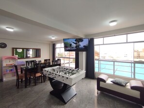 A room with a foosball table, a TV mounted on the wall, a sofa, and a window with a view of buildings.