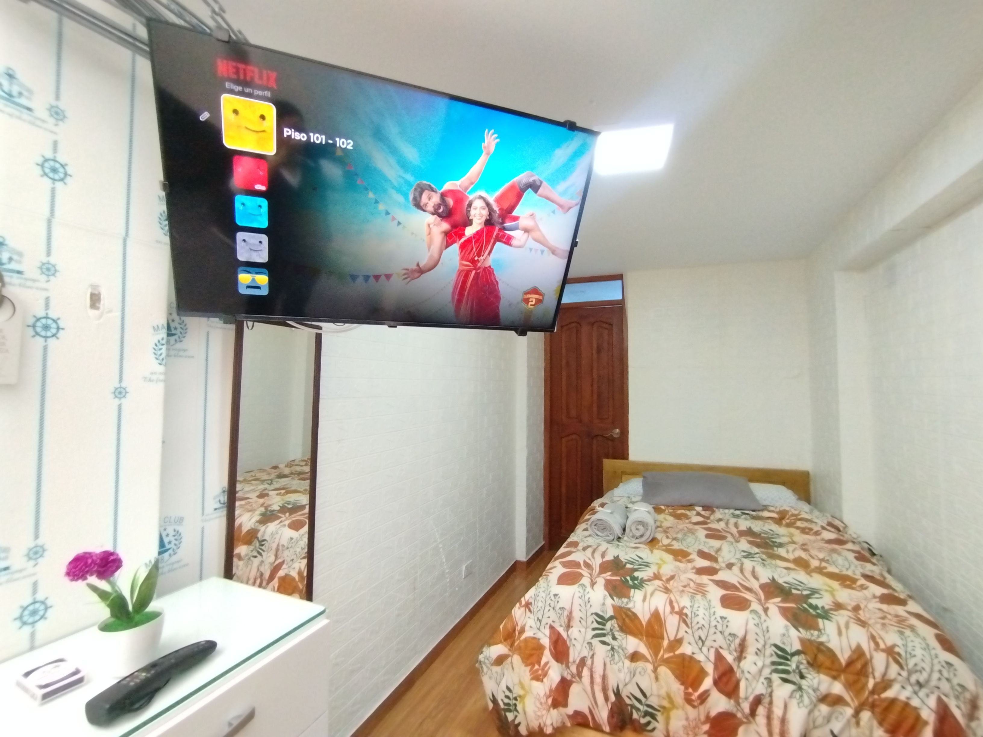 A hotel room with a television mounted on the wall, a bed with a floral bedspread, a bedside table with a remote control, and a potted plant.