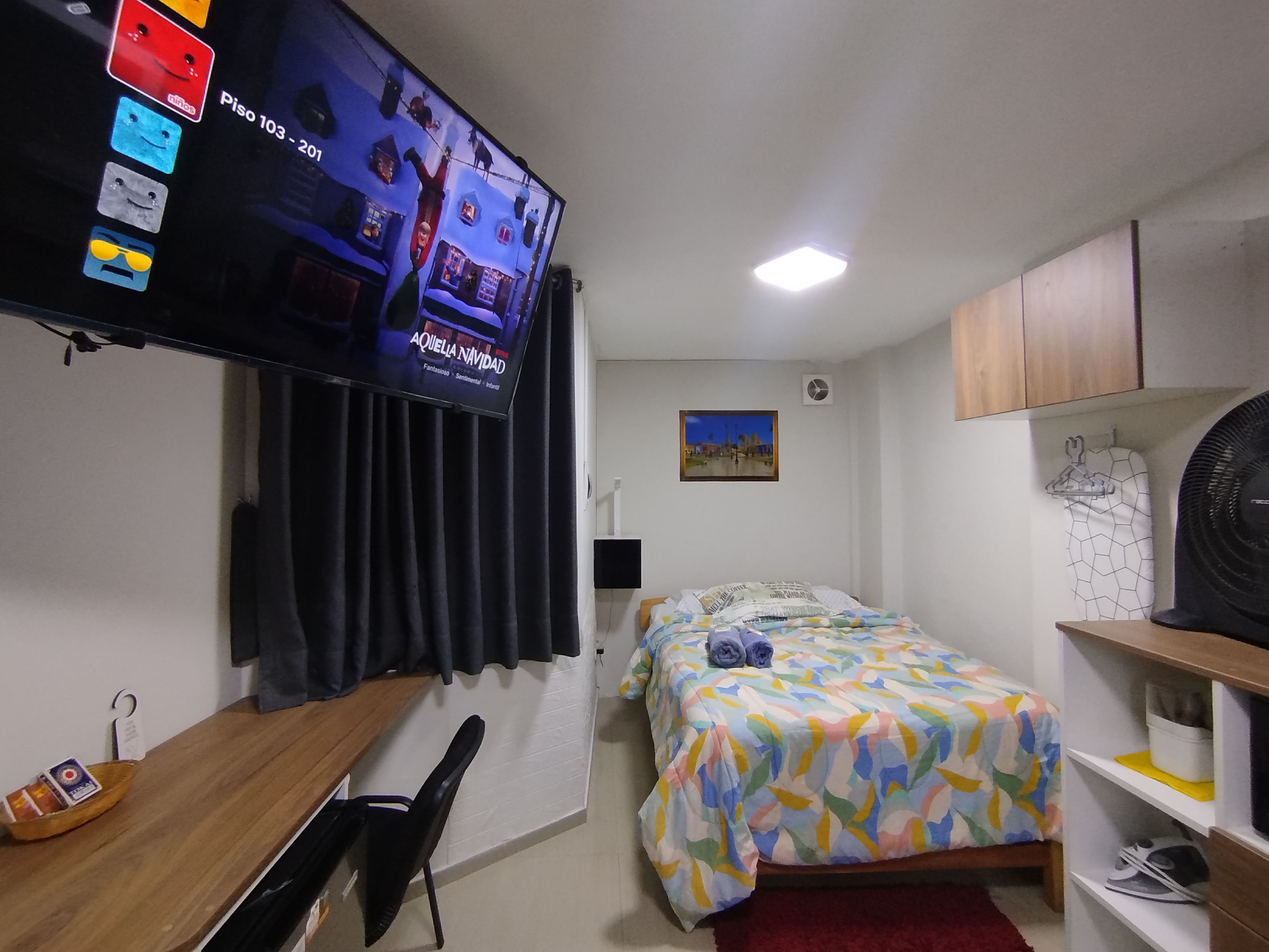 A hotel room with a bed, a television mounted on the wall, a desk with a chair, a small table, and a red rug.