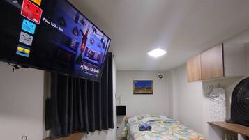 A hotel room with a bed, a television mounted on the wall, a desk with a chair, a small table, and a red rug.