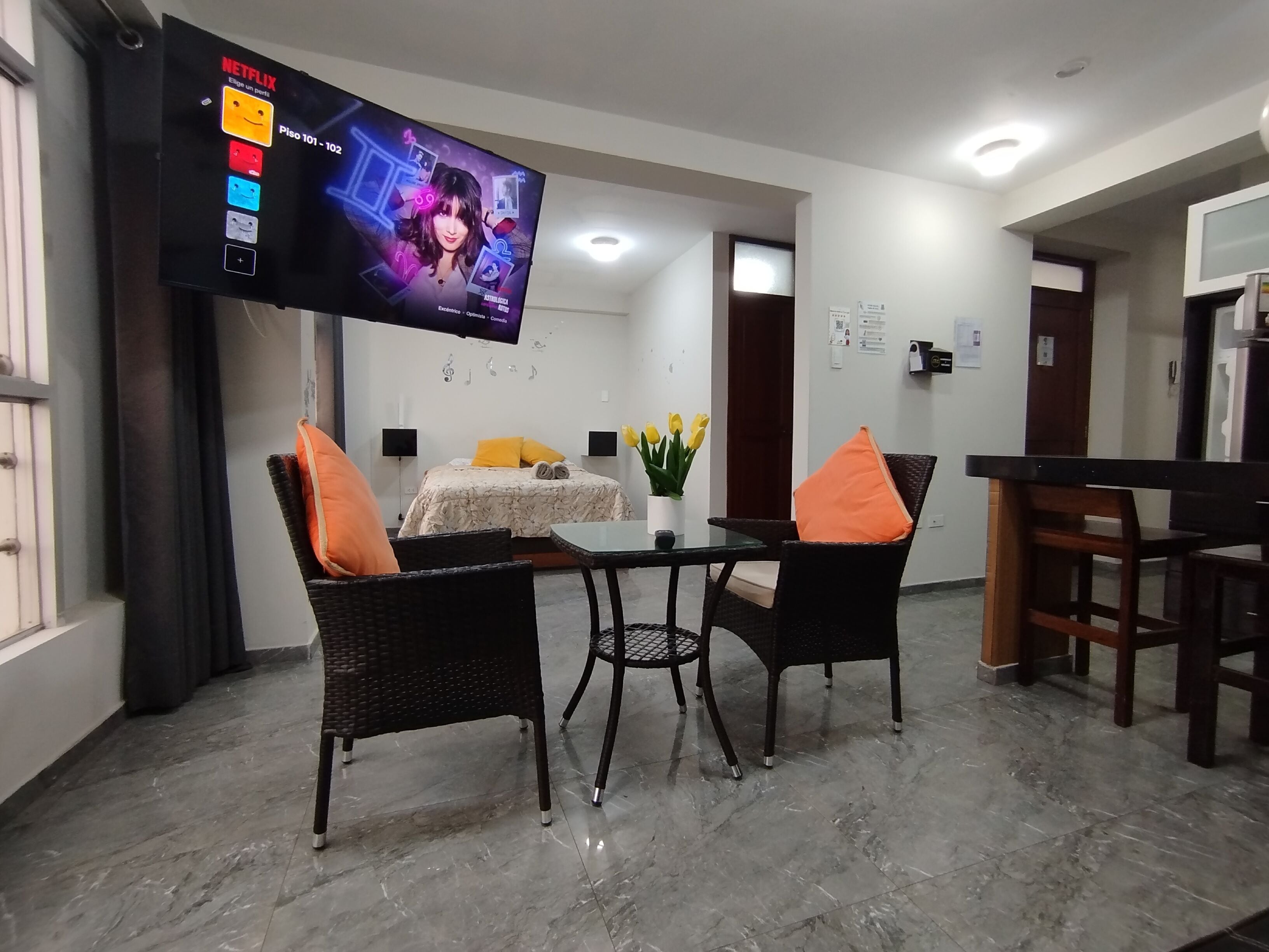 A hotel room with a bed, a TV mounted on the wall, a dining table with chairs, and a small table with a vase of flowers.