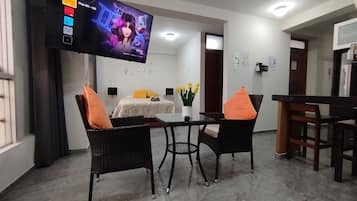 A hotel room with a bed, a TV mounted on the wall, a dining table with chairs, and a small table with a vase of flowers.