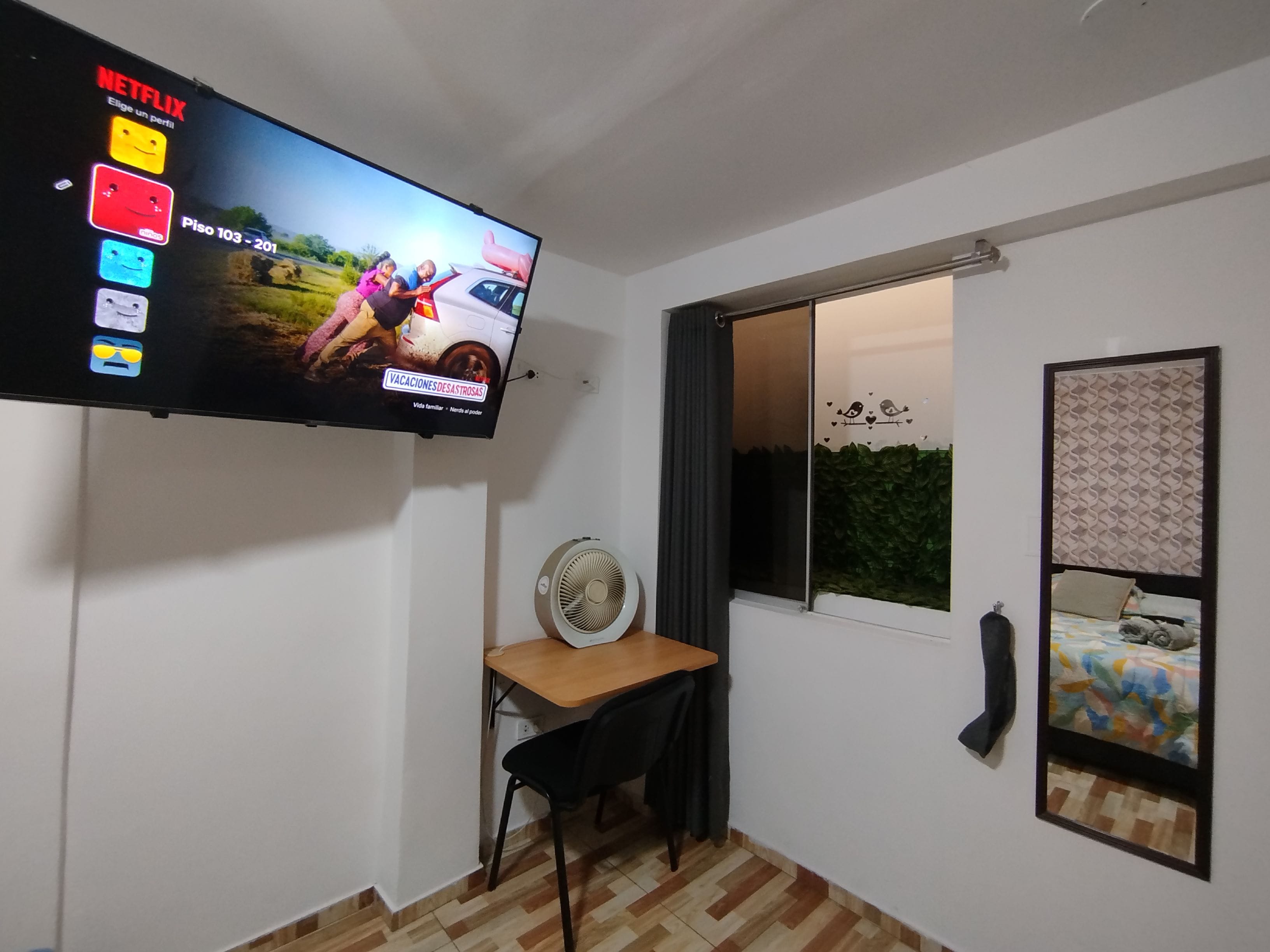 Une chambre dotée d’un téléviseur à écran plat fixé au mur, d’un bureau avec une chaise, d’une fenêtre donnant sur un paysage verdoyant et d’un lit recouvert d’une couvre-lit à motifs.