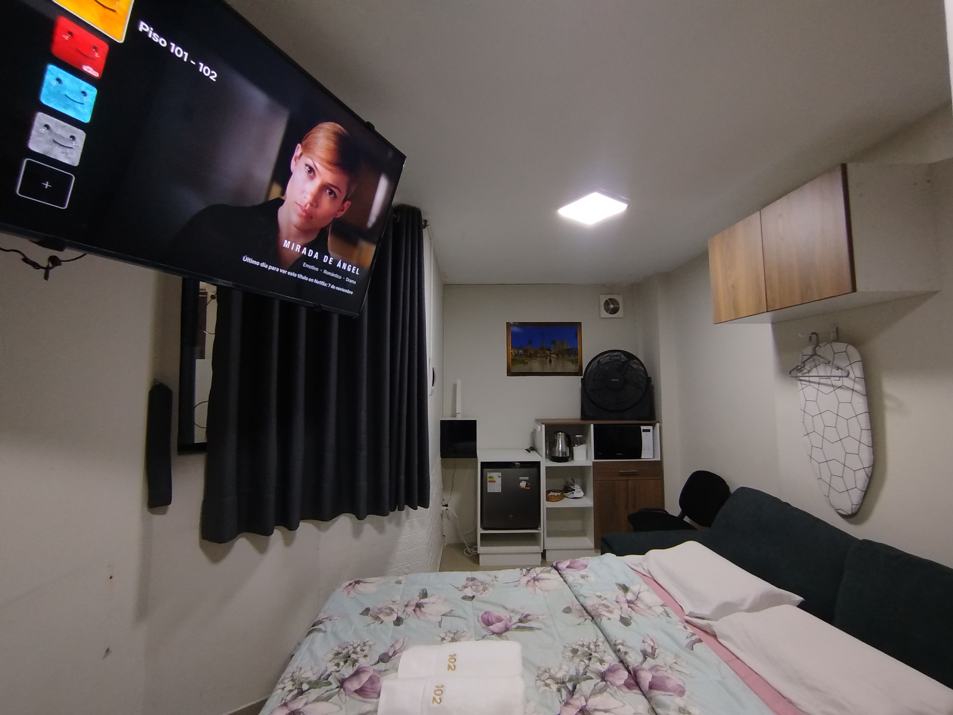 Un dormitorio con cama, televisor montado en la pared, escritorio, silla, microondas y un mueble pequeño.