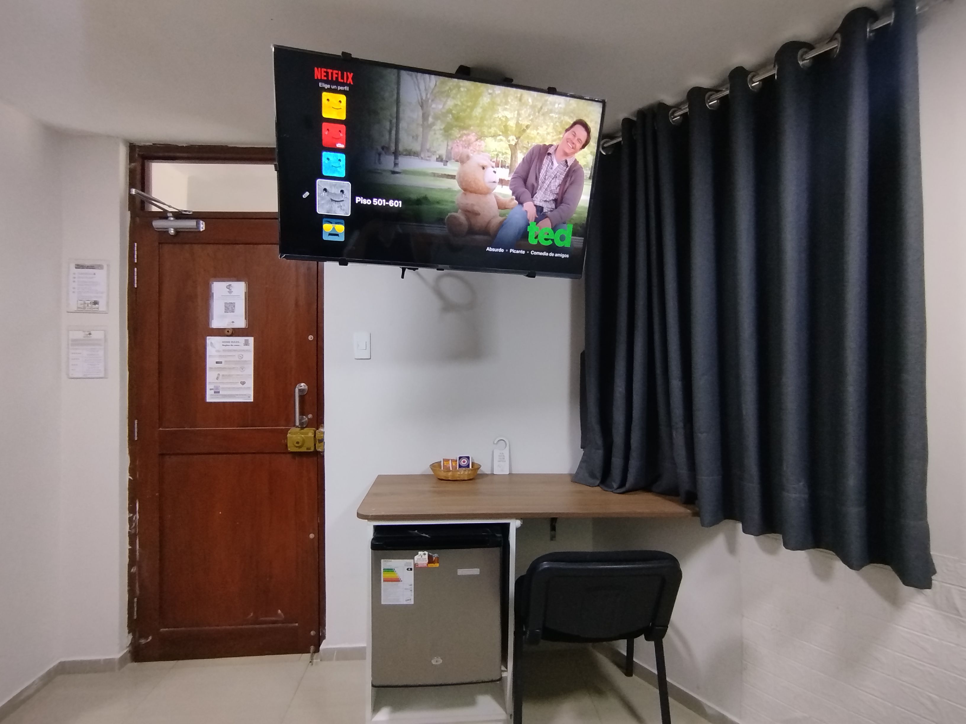 A room with a television mounted on the wall, a wooden desk, a chair, and a door with a lock.