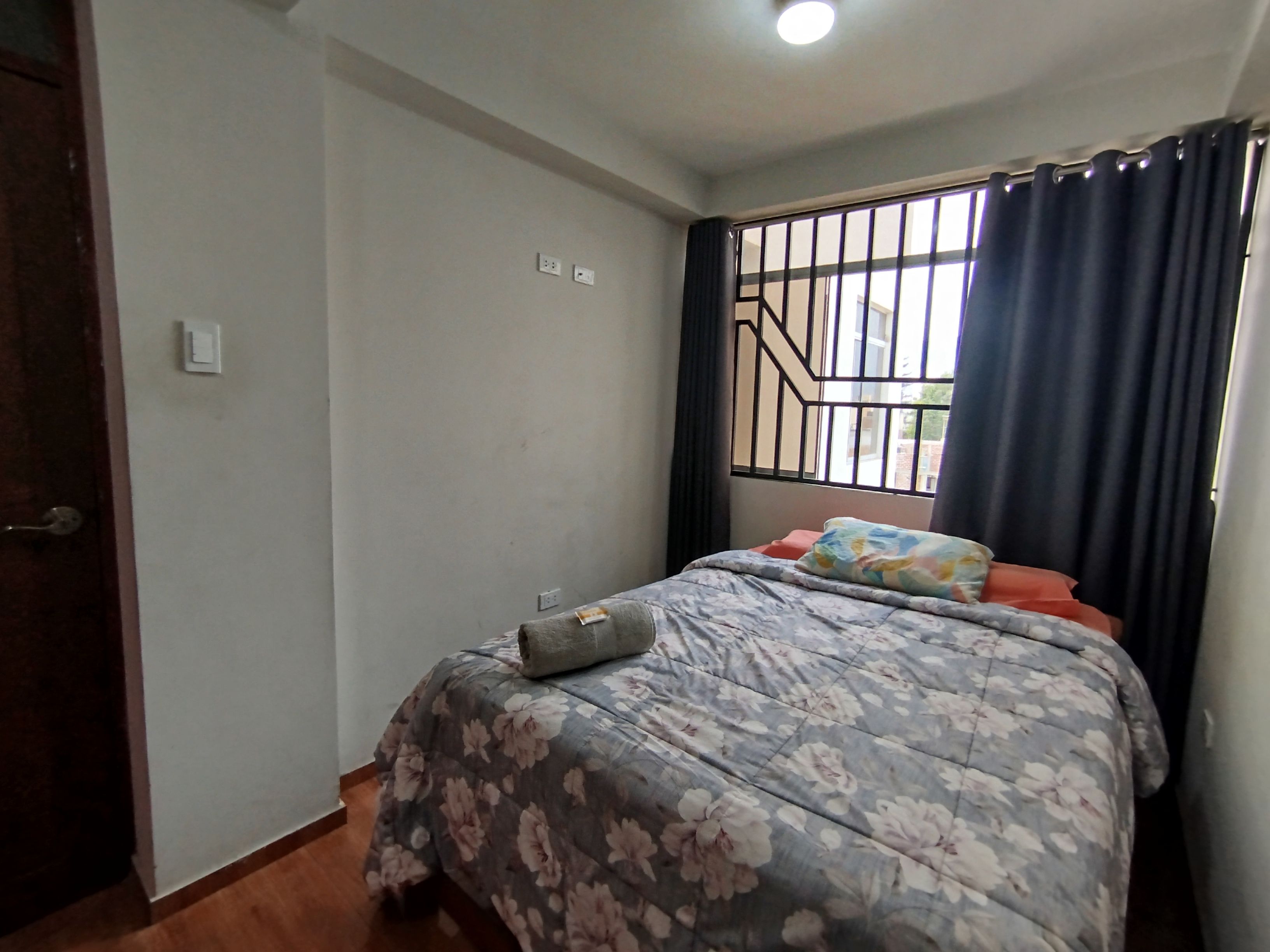 A bedroom with a bed, a window with bars, a door, and a light switch on the wall.