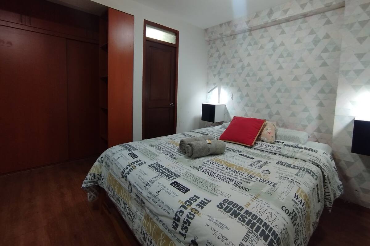 Ein Schlafzimmer mit einem Bett, das mit einem Bettbezug mit Zeitungsmotiv bezogen ist, einem roten Kissen und einem zusammengerollten Handtuch. Es gibt einen Holzschrank und eine Tür mit einer satinierten Glasscheibe.
