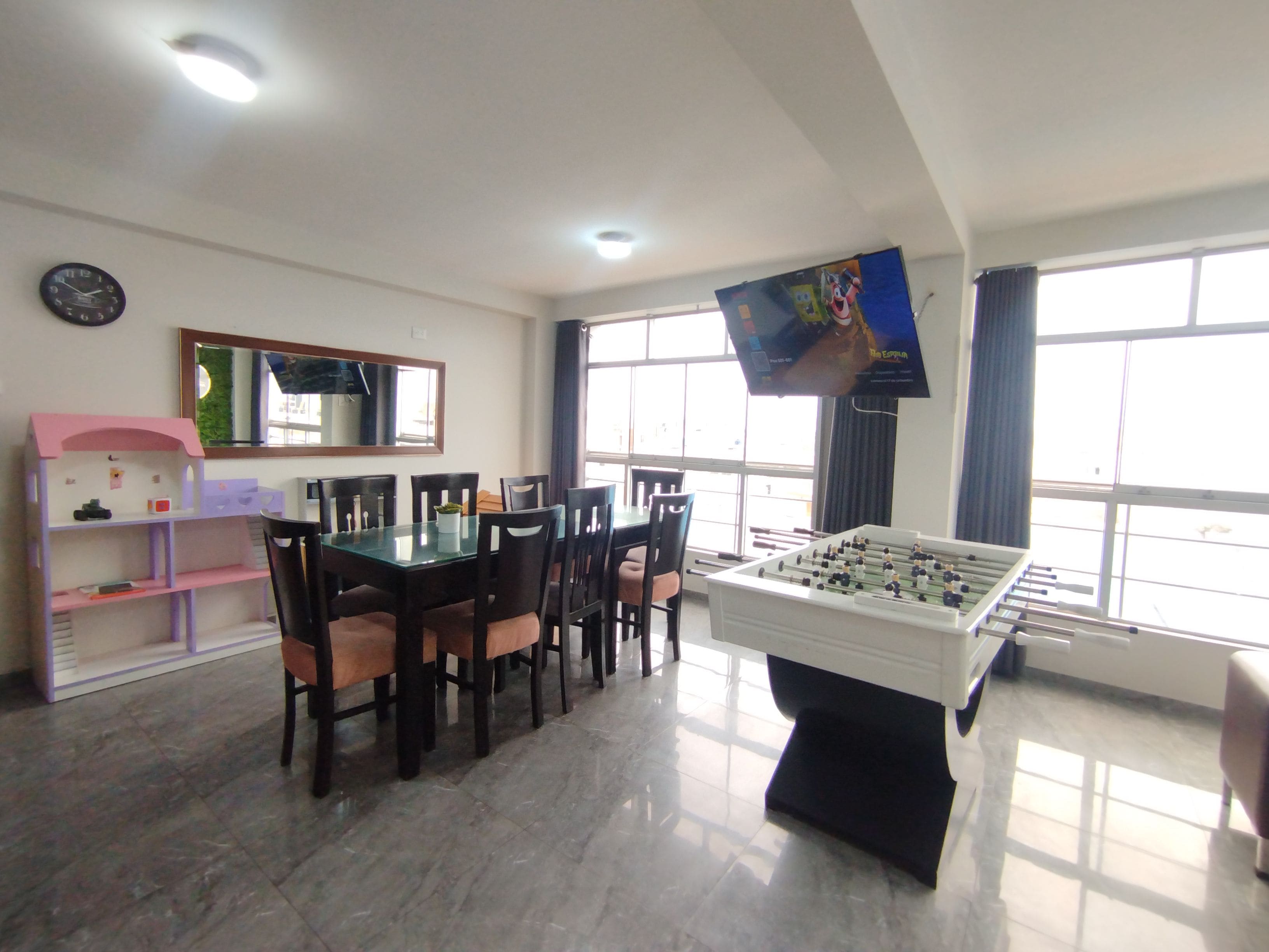 A room with a foosball table, a dining table with chairs, a TV on the wall, and a window with curtains.