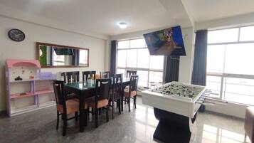 A room with a foosball table, a dining table with chairs, a TV on the wall, and a window with curtains.
