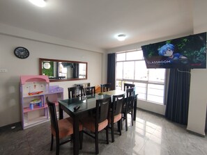 A dining area with a glass table, chairs, a television, a clock, and a window with curtains.