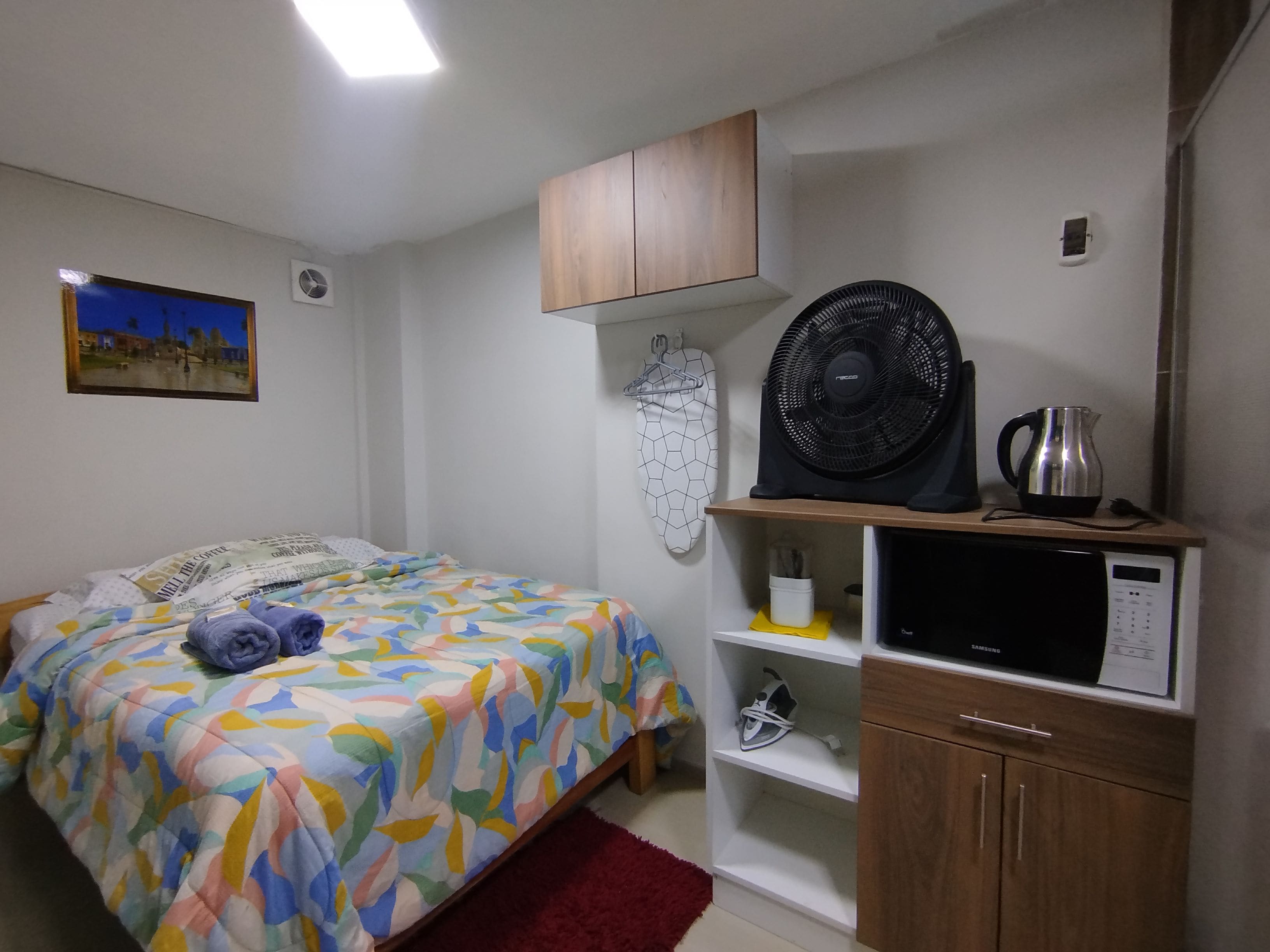 A bedroom with a bed, a fan, a microwave, and a shelf with a kettle.