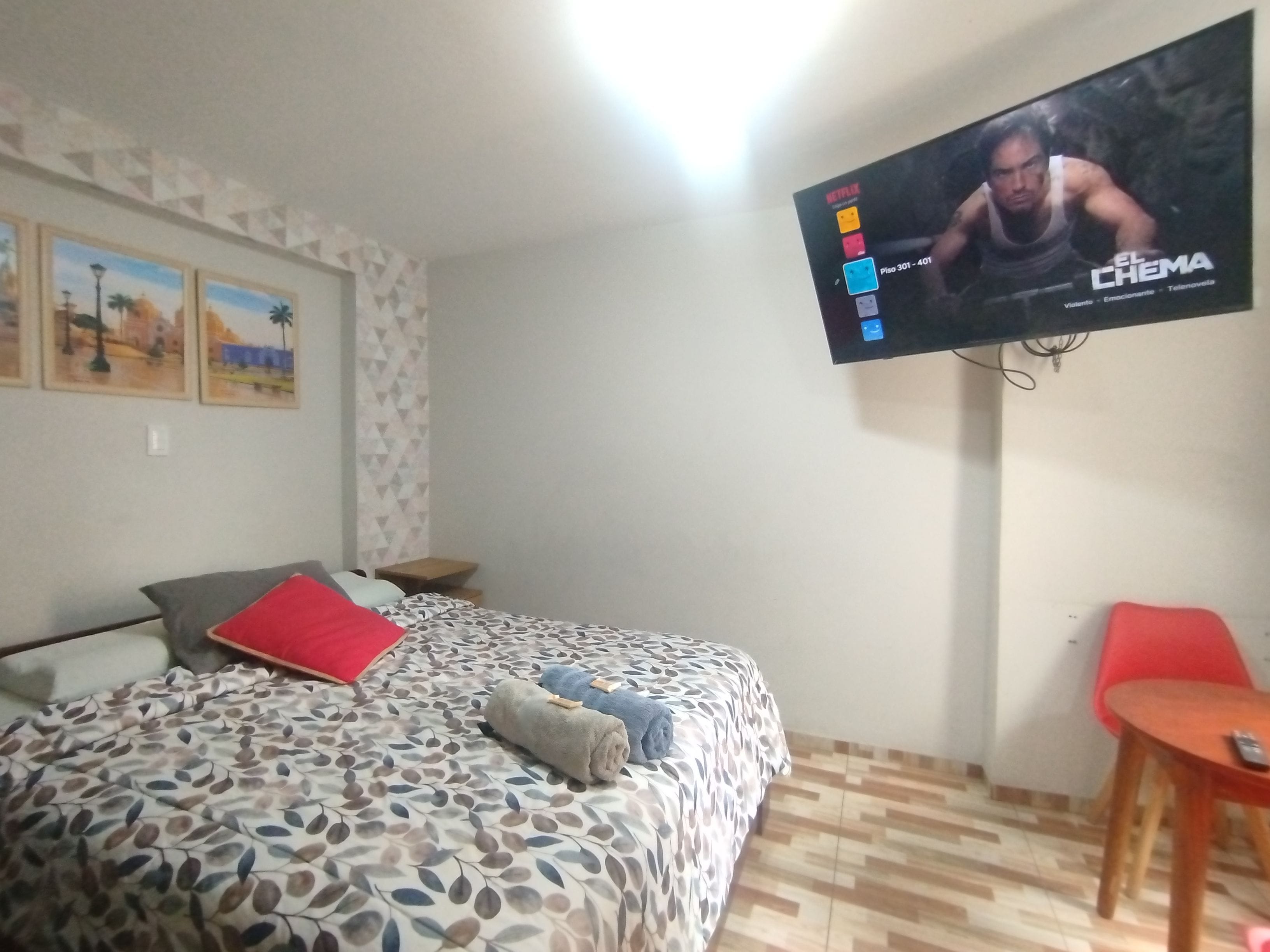 Una habitación de hotel con una cama, un televisor montado en la pared, una mesita pequeña y una silla roja.