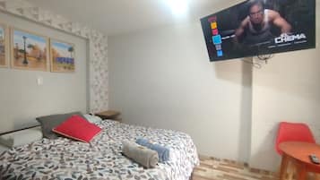 Una habitación de hotel con una cama, un televisor montado en la pared, una mesita pequeña y una silla roja.