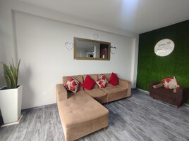 A living room with a sofa, ottoman, and a small table with a chair. There is a potted plant, a mirror, and a wall with a green accent.