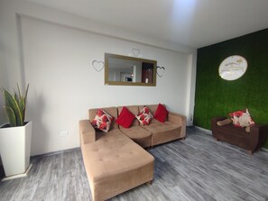 A living room with a sofa, ottoman, and a small table with a chair. There is a potted plant, a mirror, and a wall with a green accent. - Departamentos Appart d'Eliette (Trujillo)
