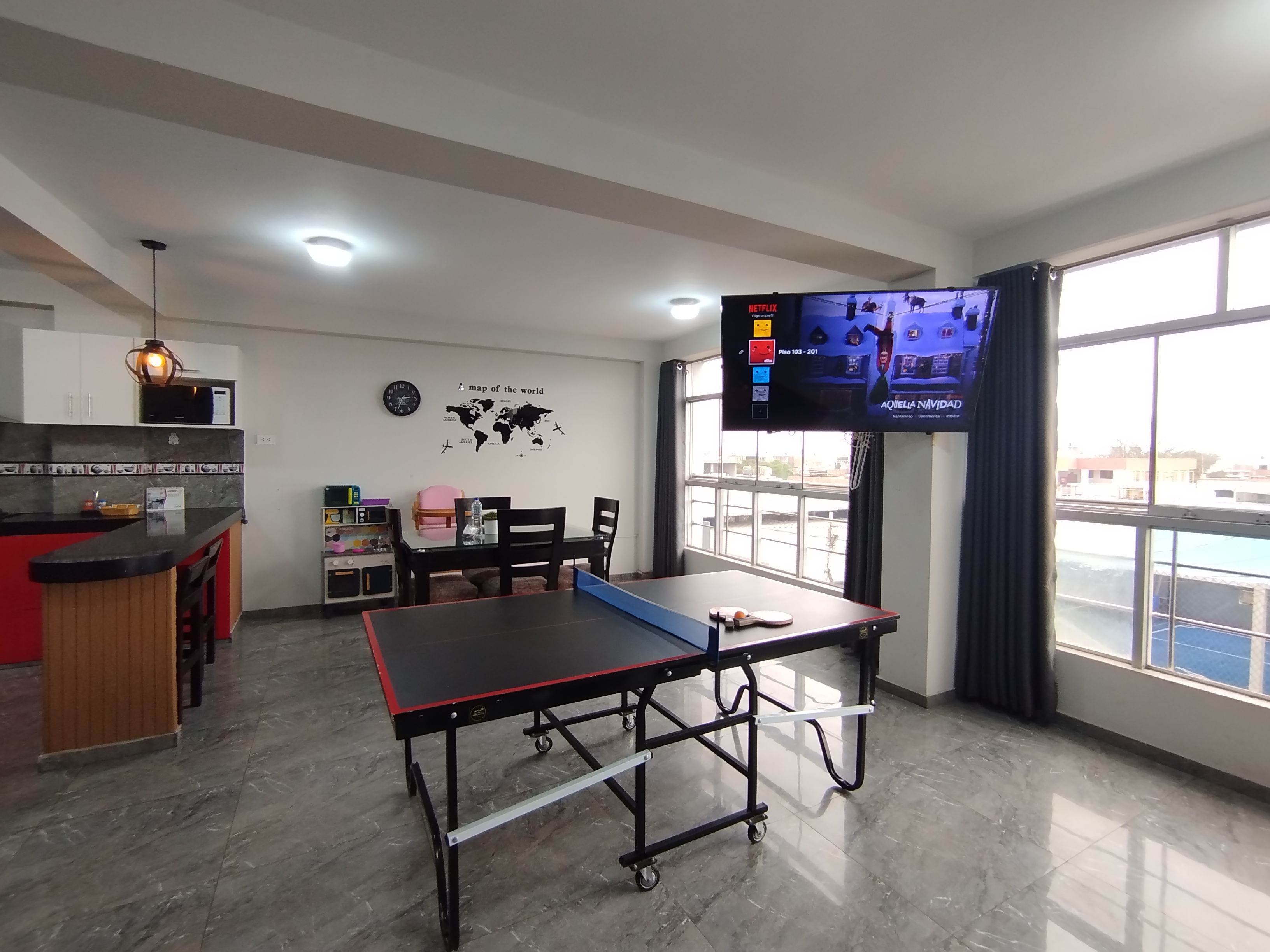 Una habitación con mesa de ping pong, comedor con sillas y mesa, cocina con microondas y televisor mostrando una película.