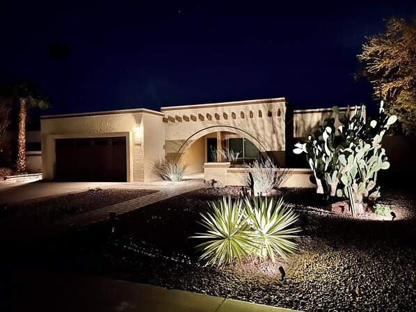 A modern house with a garage, arched entryway, and well-lit garden with cacti and agave plants.