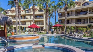 A hotel pool area with multiple levels of balconies, palm trees, and red umbrellas.