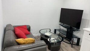 Ett modernt vardagsrum med en grå soffa, röda och gula kuddar, ett soffbord i glas, ett svart TV-bord och ett vitt skåp.