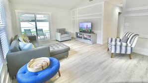 Ein geräumiges Wohnzimmer mit einem grauen Sofa, einem blauen Hocker, einem gestreiften Sessel, einem Fernseher und einem Bücherregal.