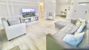 Ein modernes Wohnzimmer mit einem Flachbildfernseher, grauen Sofas, einem gestreiften Sessel und einem Essbereich im Hintergrund.