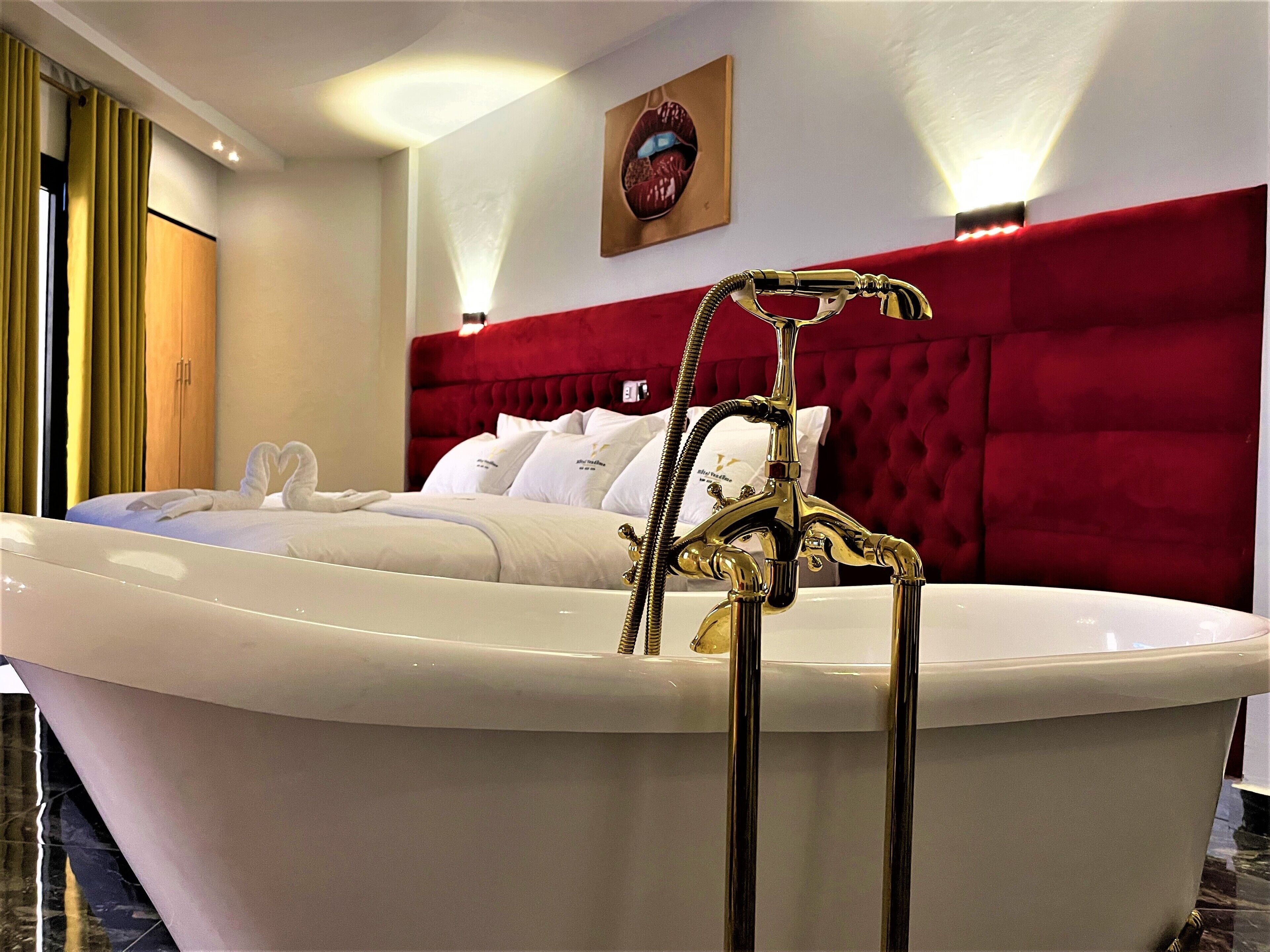 A luxurious bedroom with a large bathtub, a red upholstered headboard, and a painting of lips on the wall.