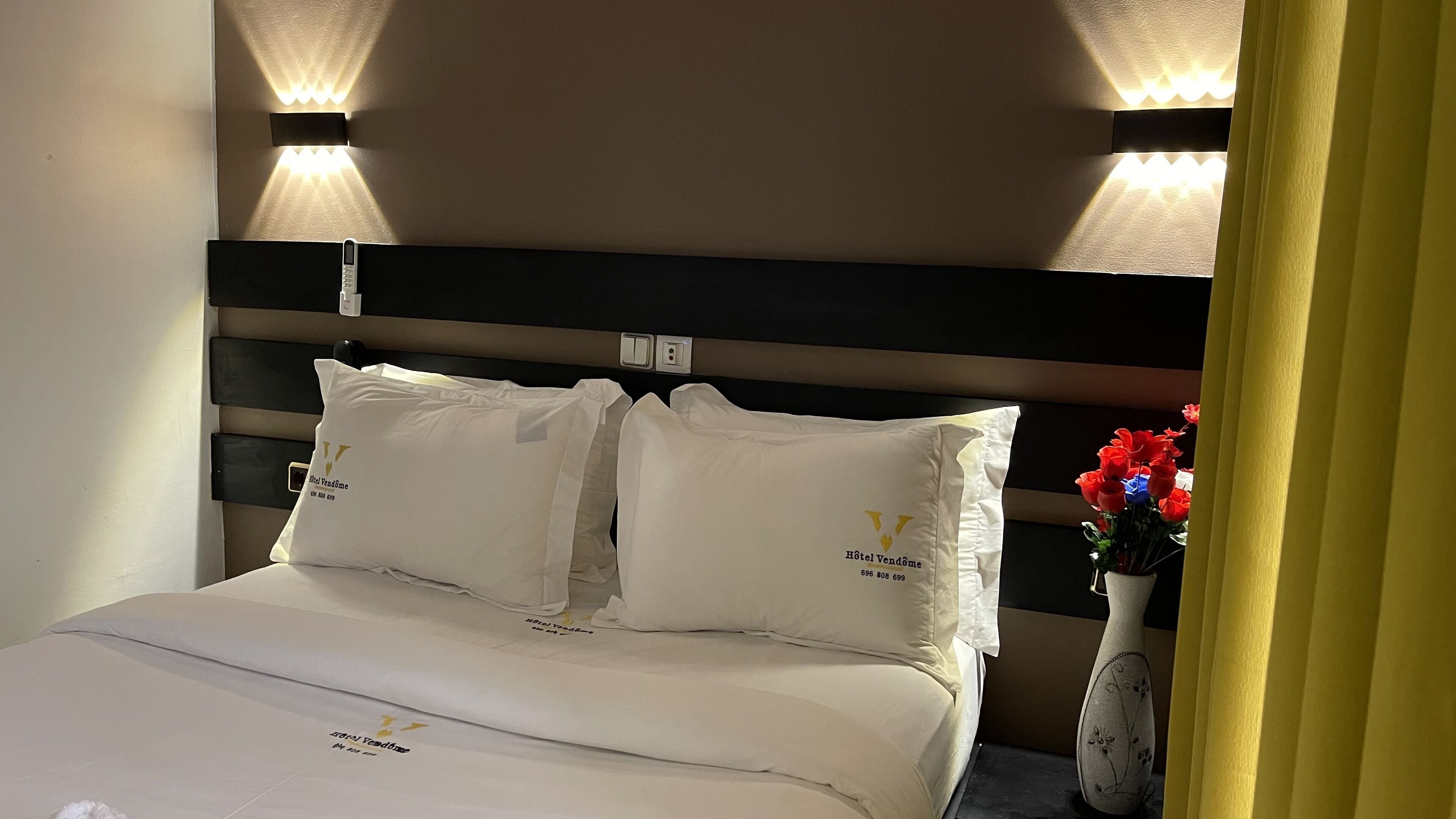 Una habitación de hotel con una cama bien hecha, dos almohadas con el logotipo del hotel, un jarrón con flores e iluminación empotrada en la pared.