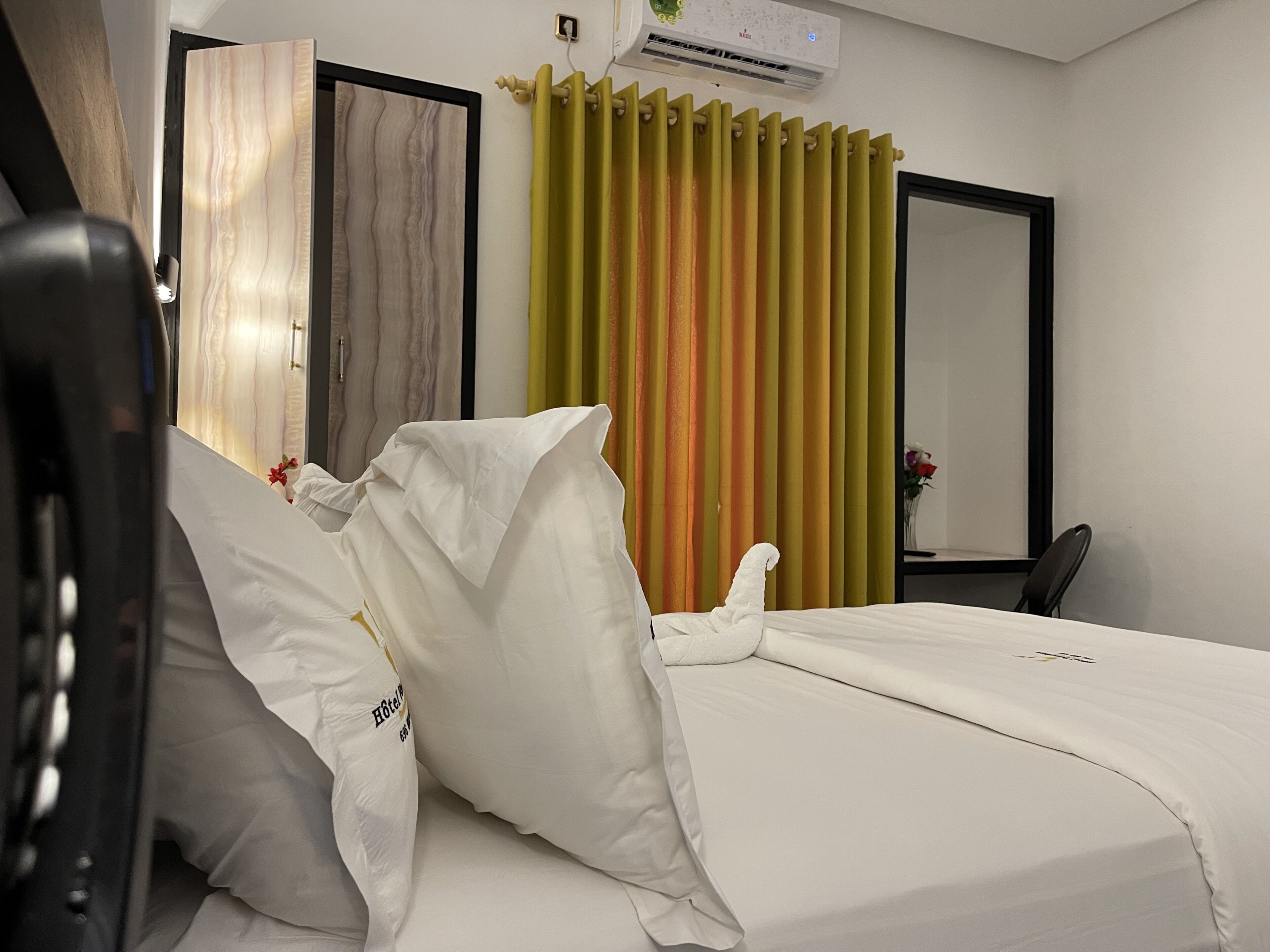 A hotel room with a bed, pillows, a chair, a mirror, and a window with curtains.