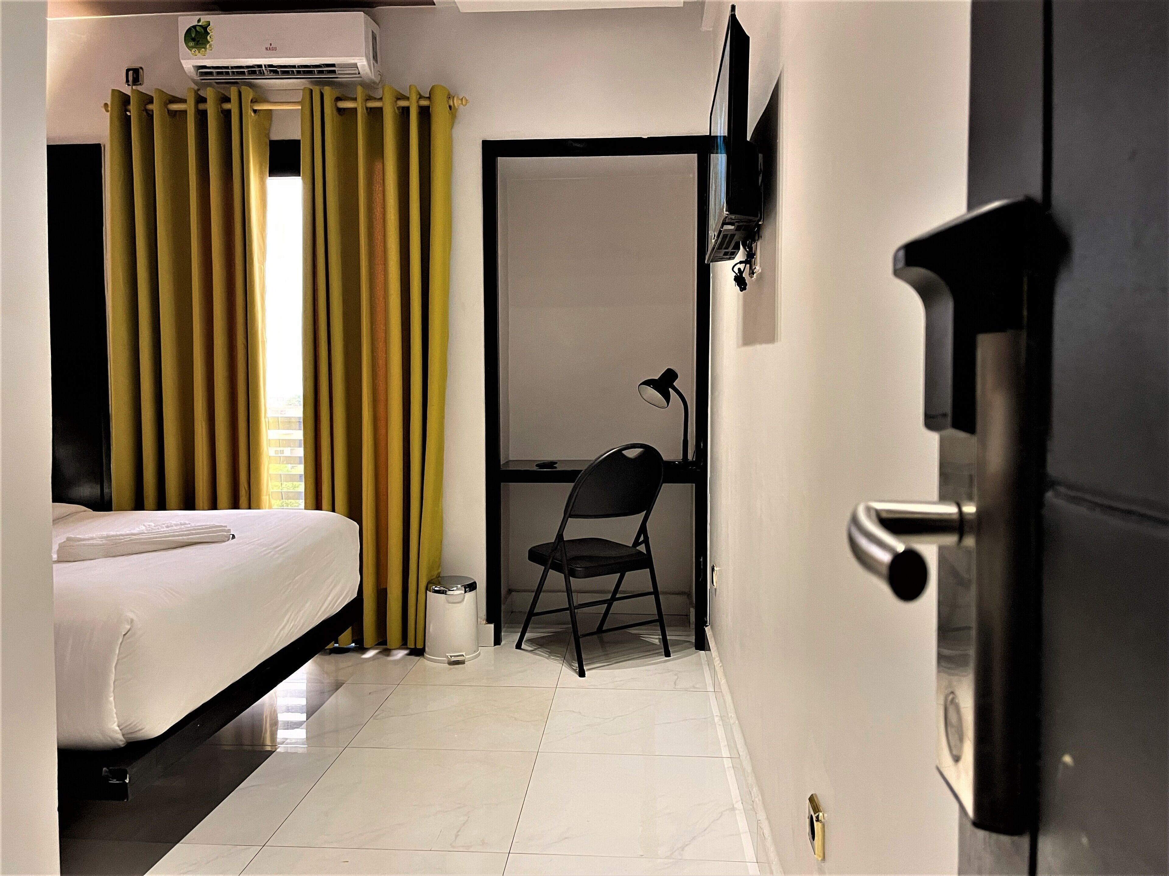 Ein Hotelzimmer mit einem Bett, einem Schreibtisch mit Stuhl, einer Schreibtischlampe, einem Papierkorb und einer Klimaanlage.