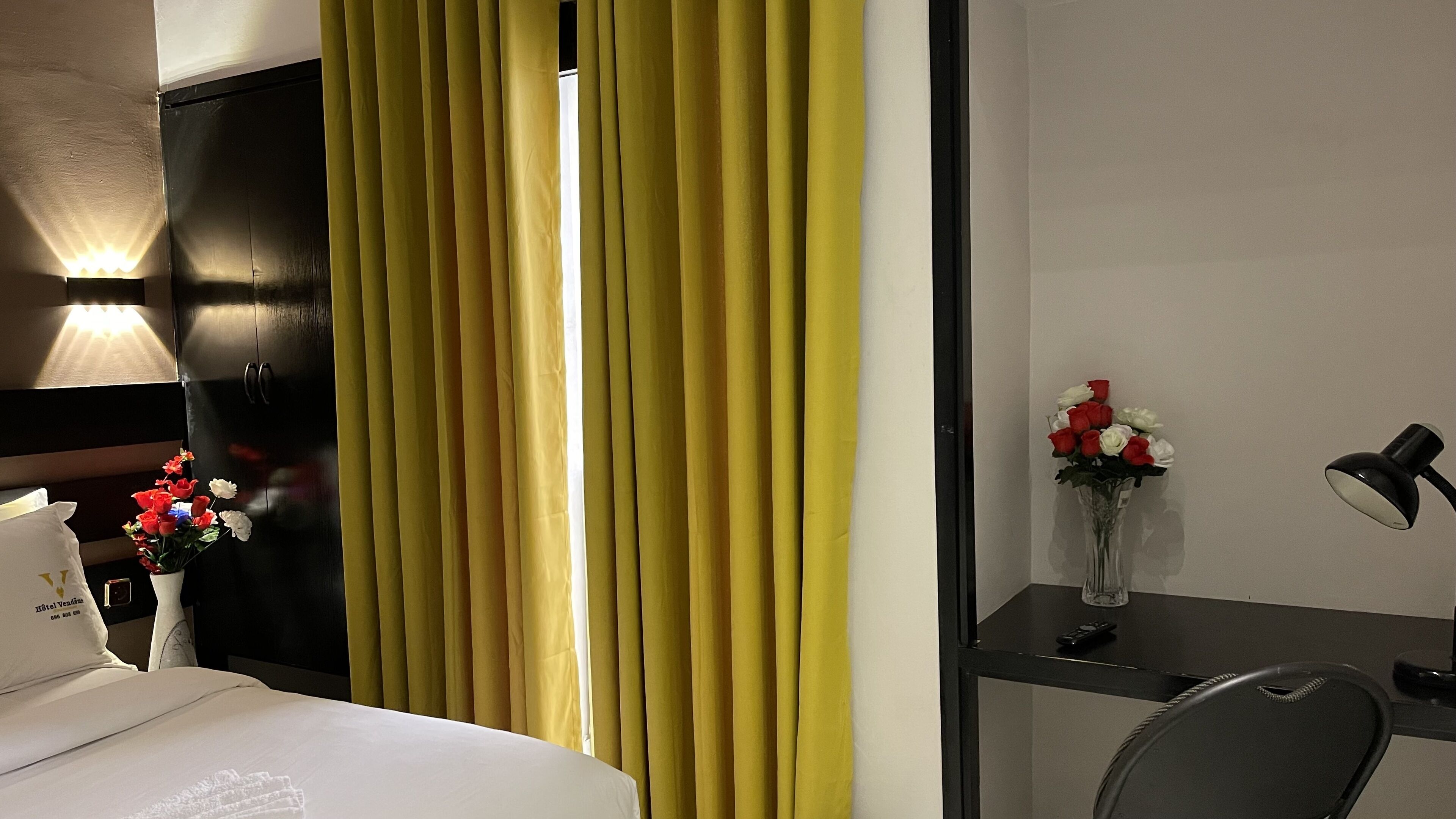 A hotel room with a bed, a desk with a lamp, a vase with flowers, and a curtain.