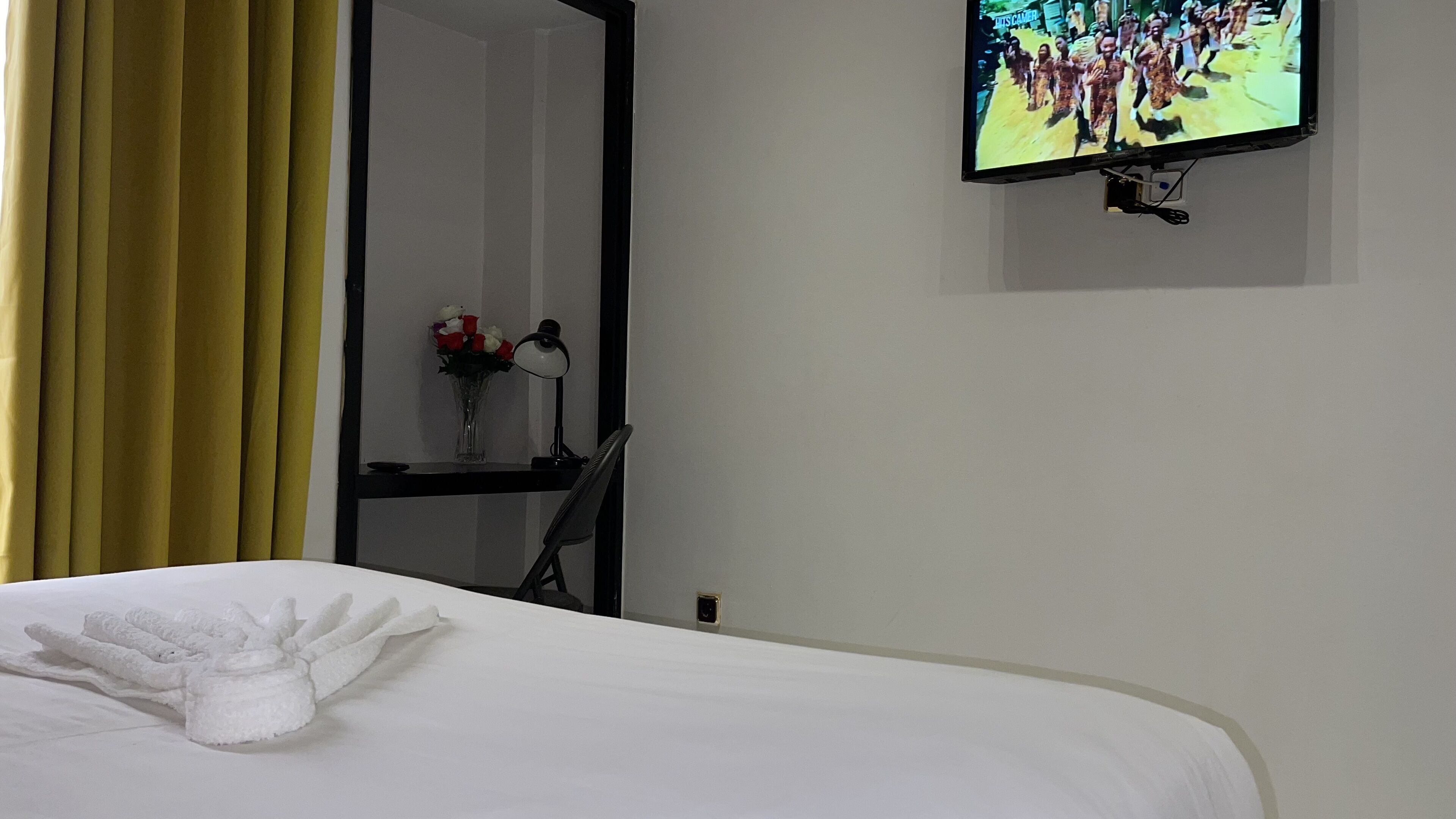 A hotel room with a bed, a TV mounted on the wall, a desk with a lamp, and a vase with flowers.