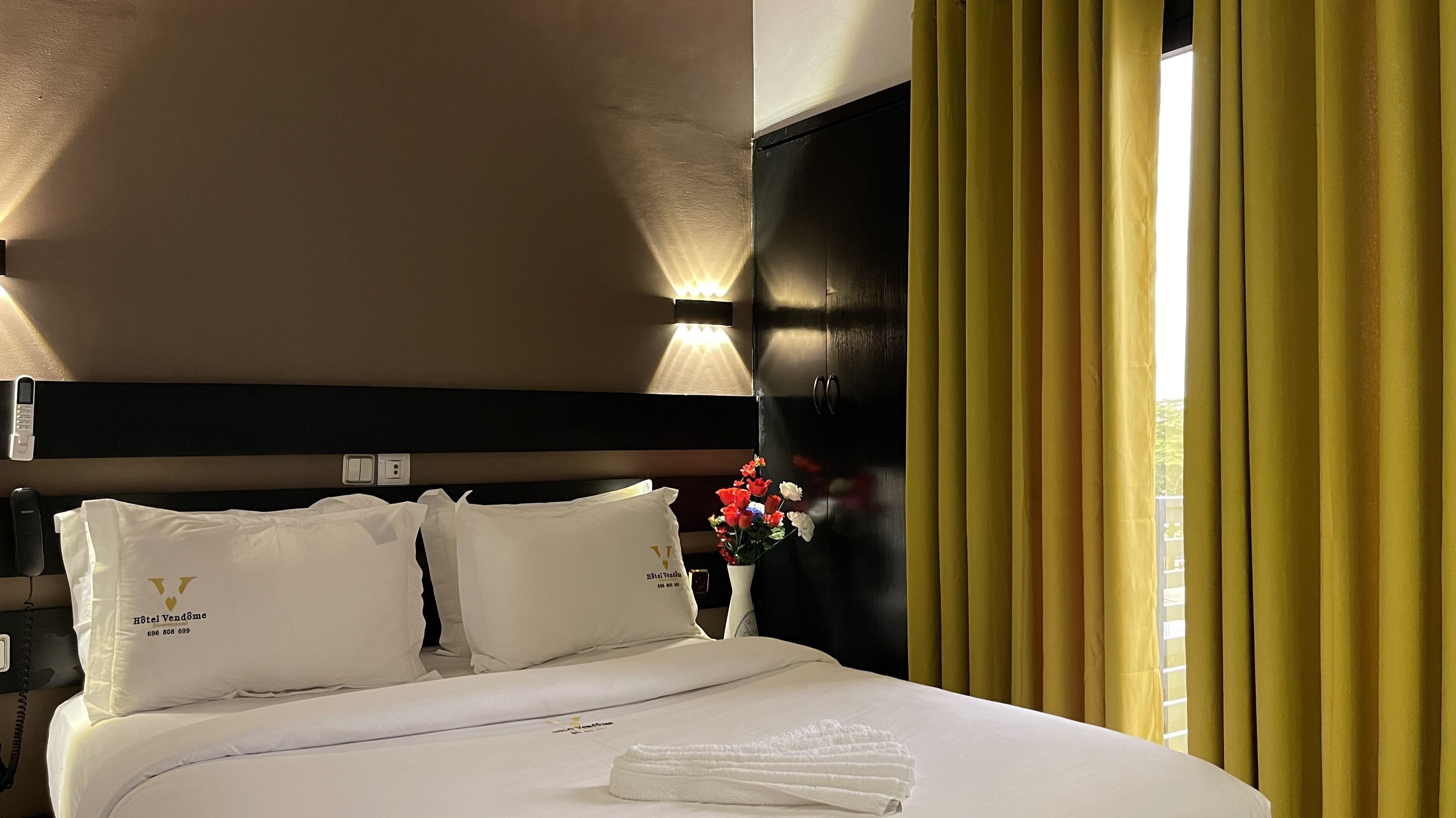 Une chambre d’hôtel avec un lit, des oreillers, une table de chevet ornée d’un vase de fleurs et une fenêtre avec des rideaux jaunes.