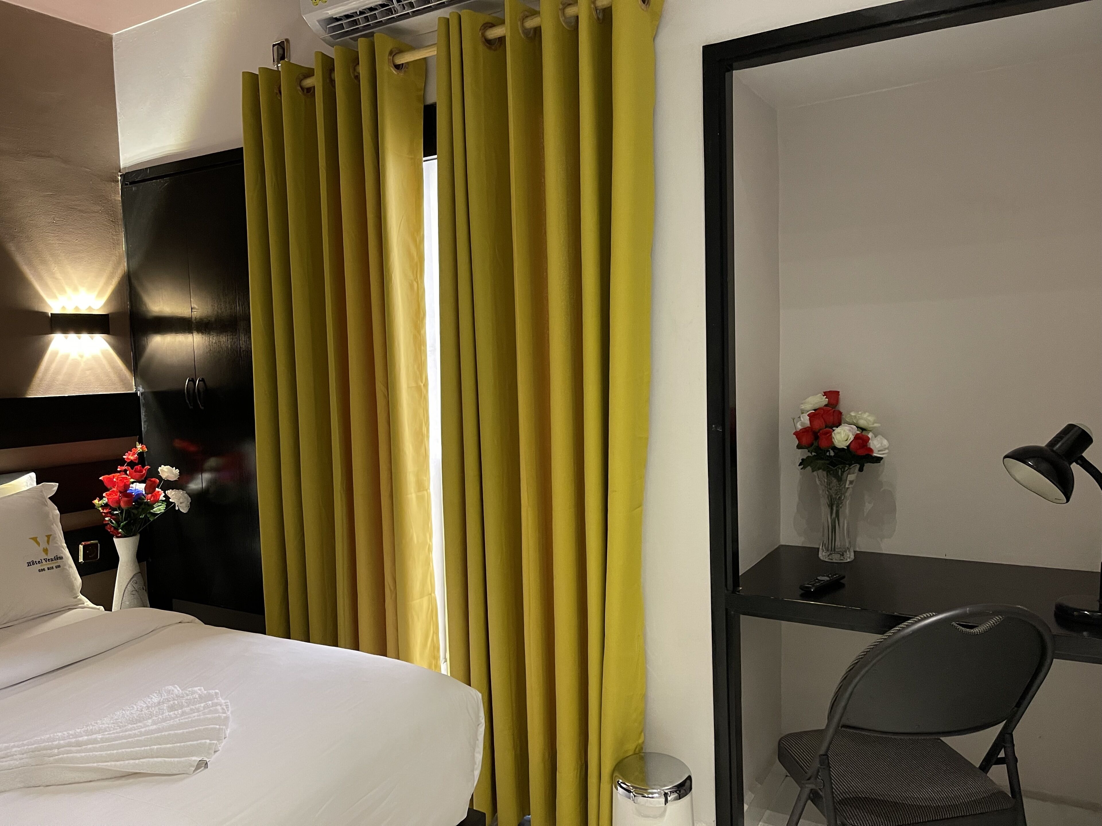 A hotel room with a bed, a desk with a chair, a trash bin, a vase with flowers, and curtains.