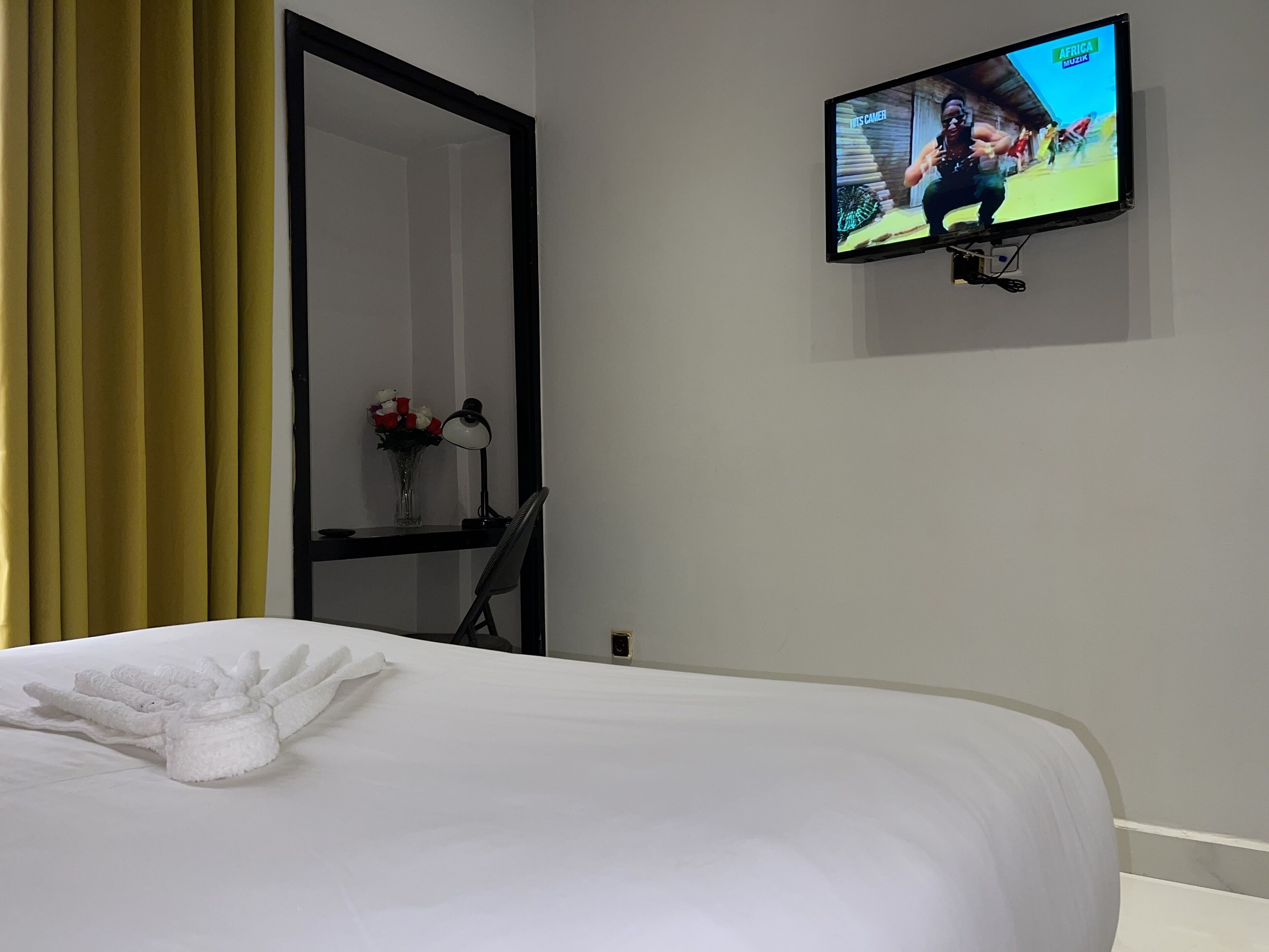 A hotel room with a bed, a TV mounted on the wall, a desk with a lamp, and a vase with flowers.