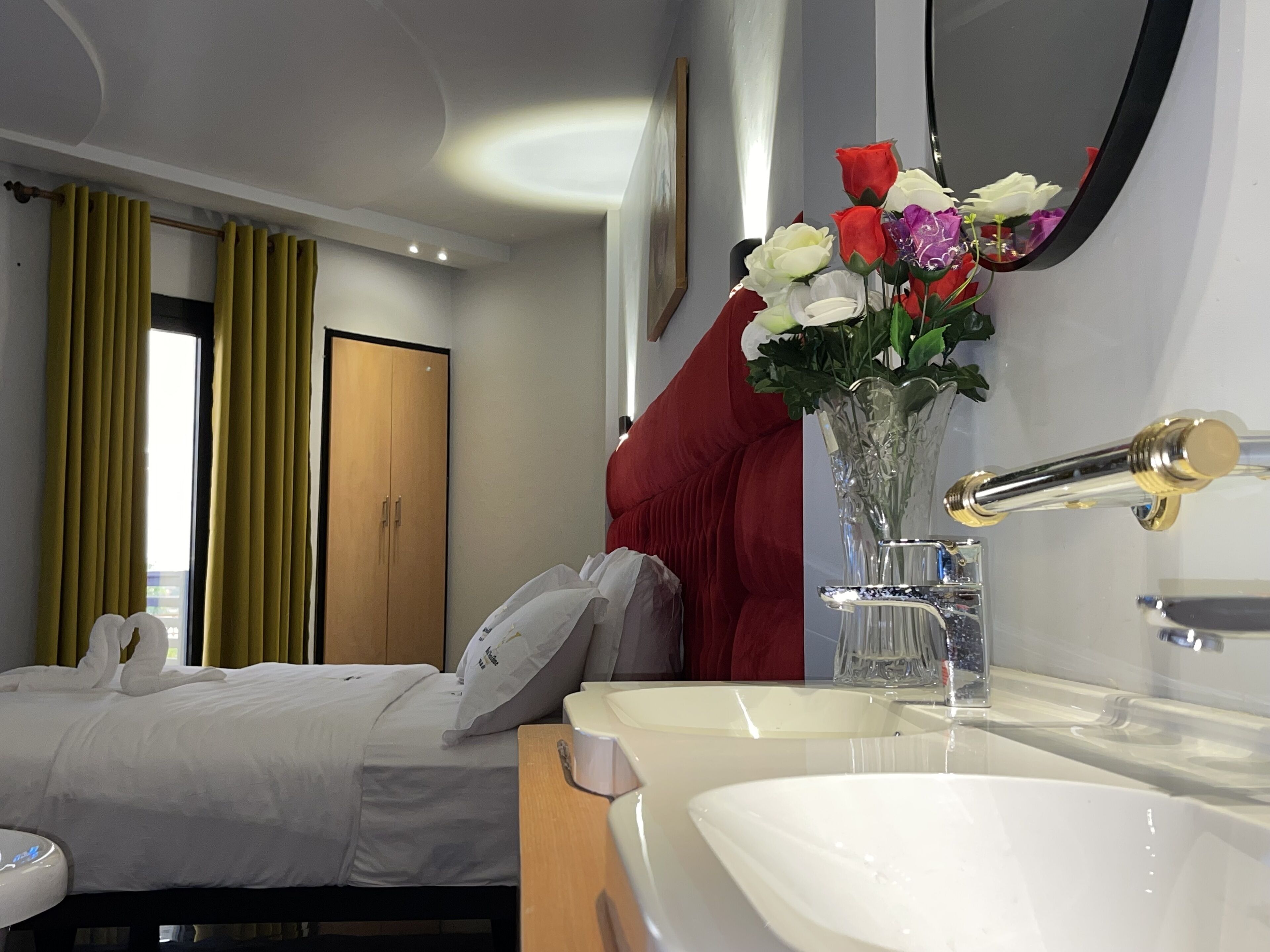 A hotel room with a bed, a bedside table, a mirror, a vase with flowers, and a window with curtains.