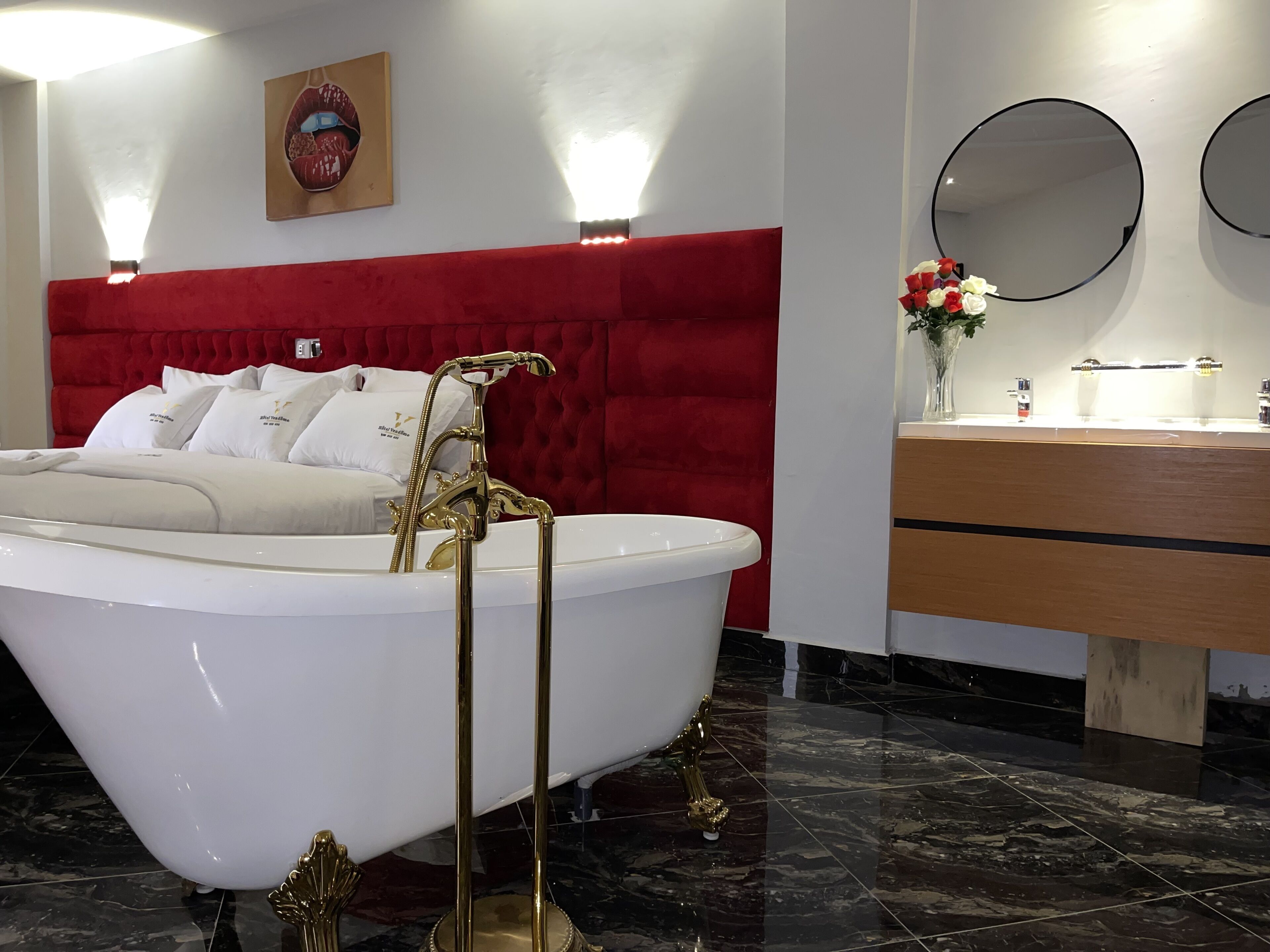 A modern bathroom with a freestanding bathtub, a red upholstered headboard, a vanity with a mirror, and a decorative wall piece.