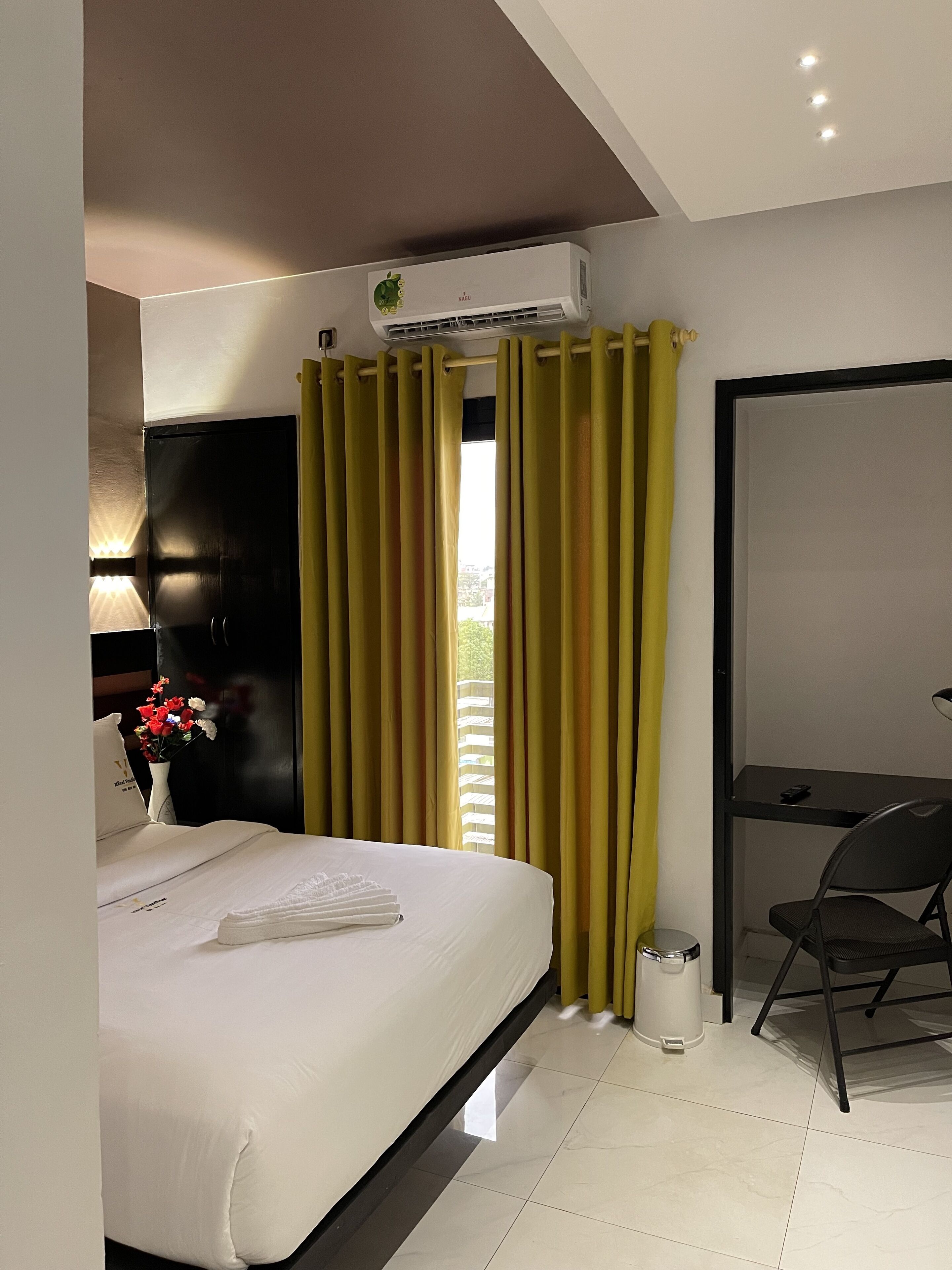 A hotel room with a bed, a desk, a chair, a trash bin, and a window with curtains.